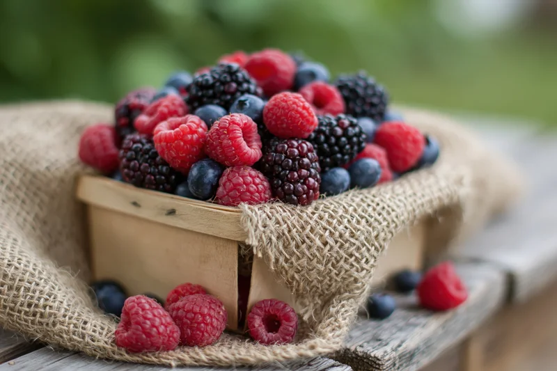 Wooden basket overflowing with fresh raspberries, blueberries and blackberries on burlap cloth outdoors
