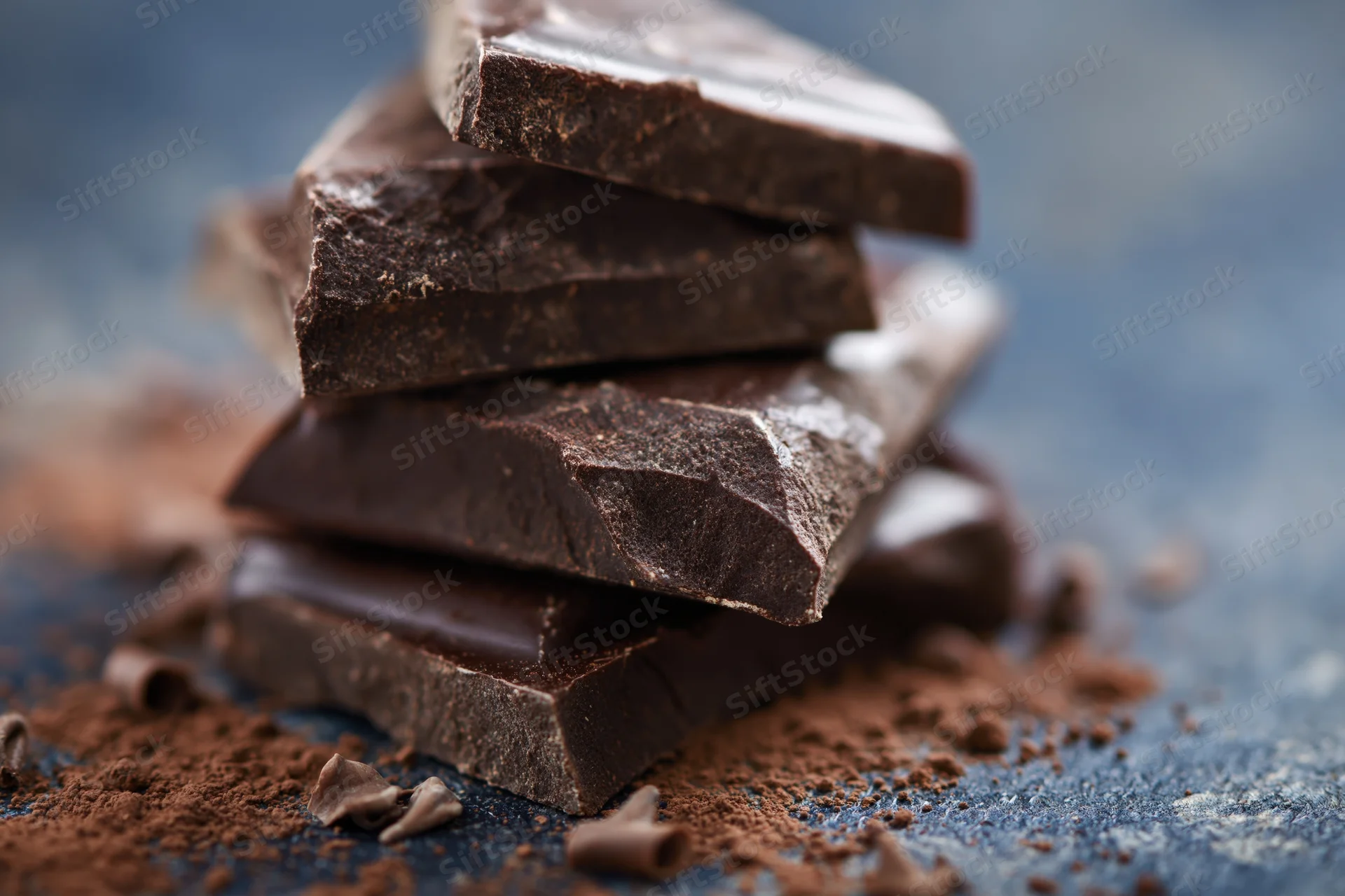 Broken dark chocolate pieces stacked loosely on a blue slate surface with cocoa powder scattered around