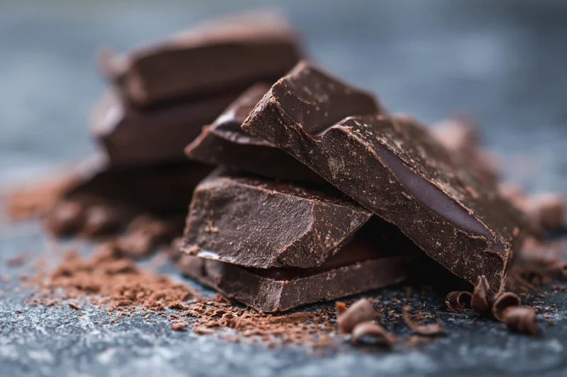 Broken dark chocolate pieces stacked loosely on a dark stone surface with cocoa powder and shavings scattered around