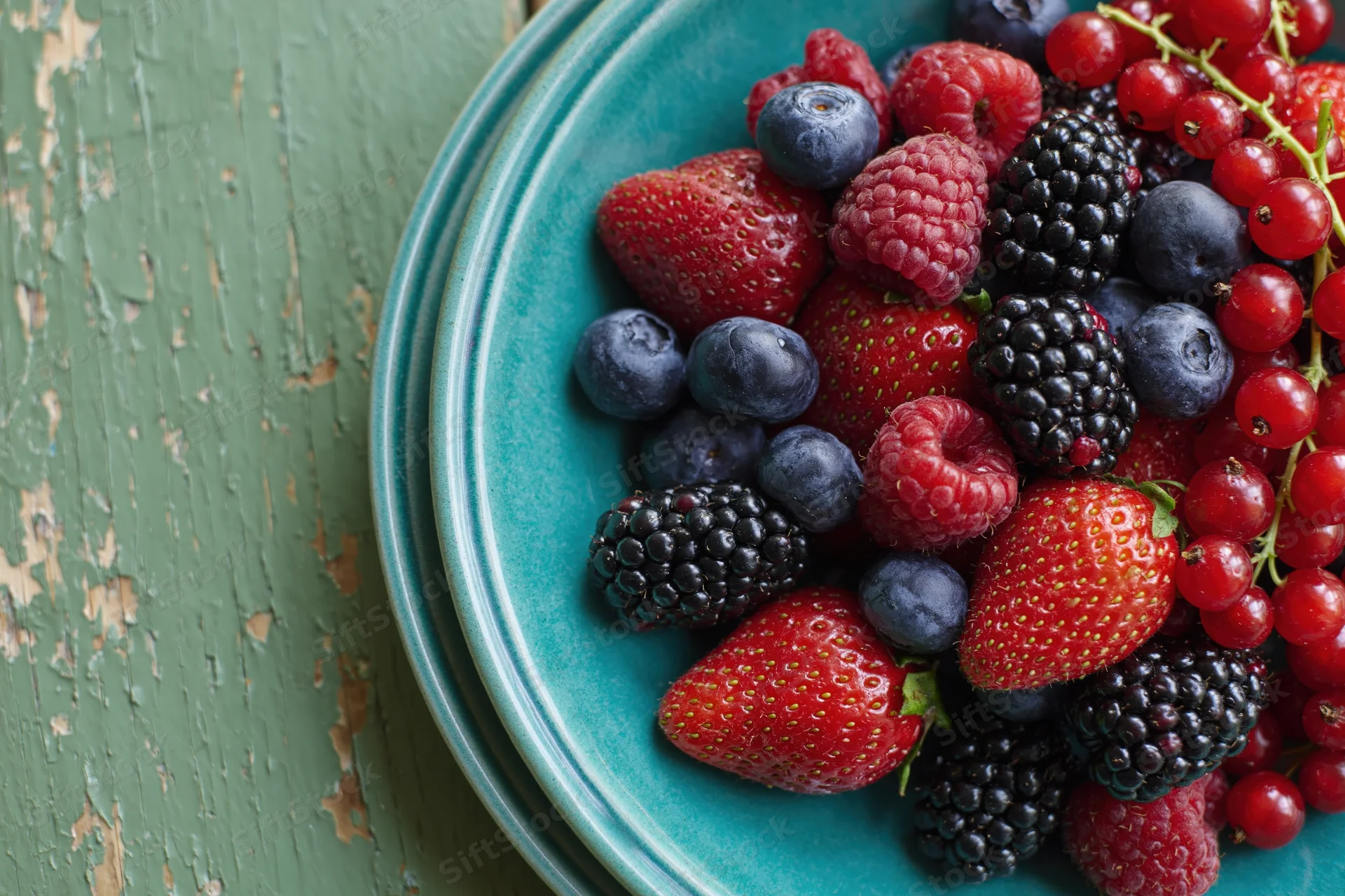 Mixed fresh berries on a teal ceramic plate on a rustic green wooden surface