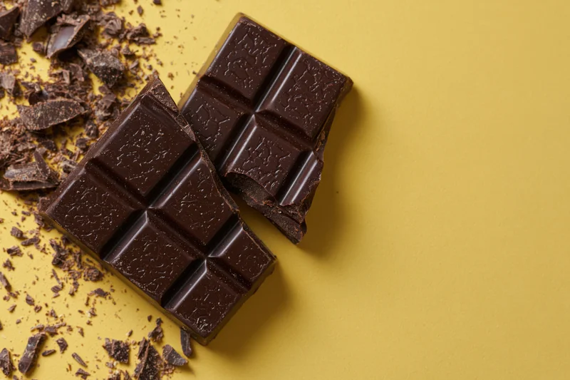 Broken dark chocolate bar pieces and crumbs on a flat yellow surface