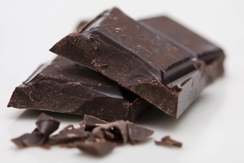 Broken dark chocolate bar pieces stacked on white surface with crumbs scattered nearby