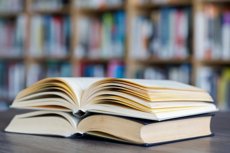 Two open hardcover books stacked on a dark wooden table in a library with blurred bookshelves in the background