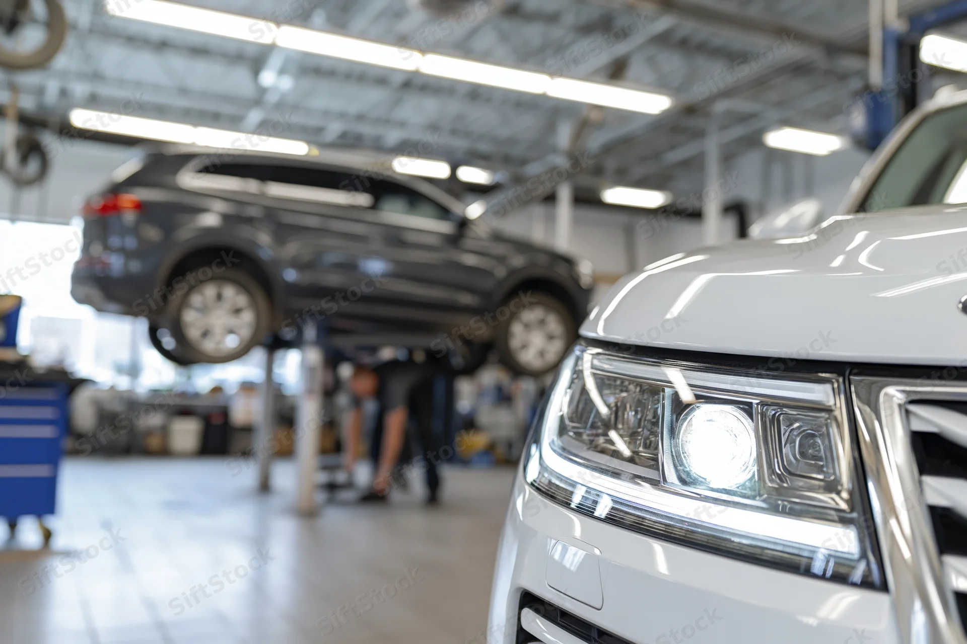 White suv front end in focus with headlight illuminated in auto repair garage, mechanic working under lifted vehicle in background