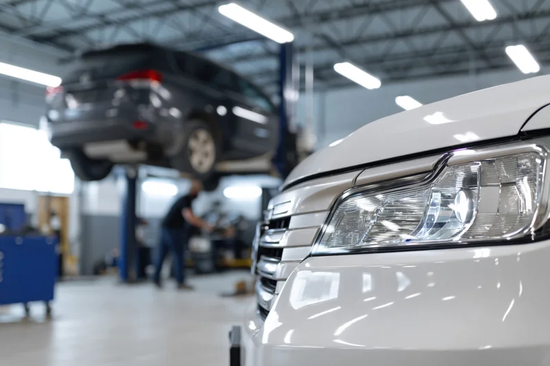 White suv front end in focus with headlight and grille in foreground, mechanic working on elevated vehicle in background garage