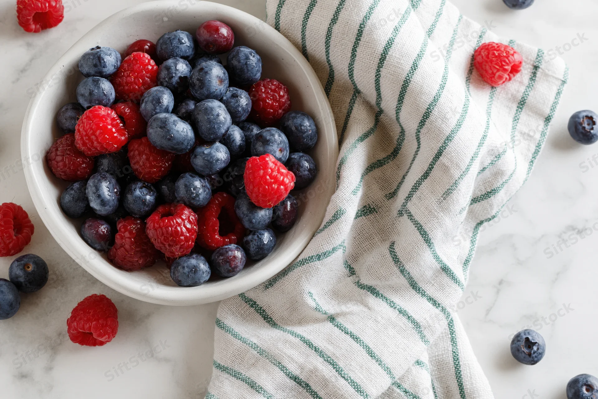 White ceramic bowl filled with fresh blueberries and raspberries on marble countertop beside striped cloth