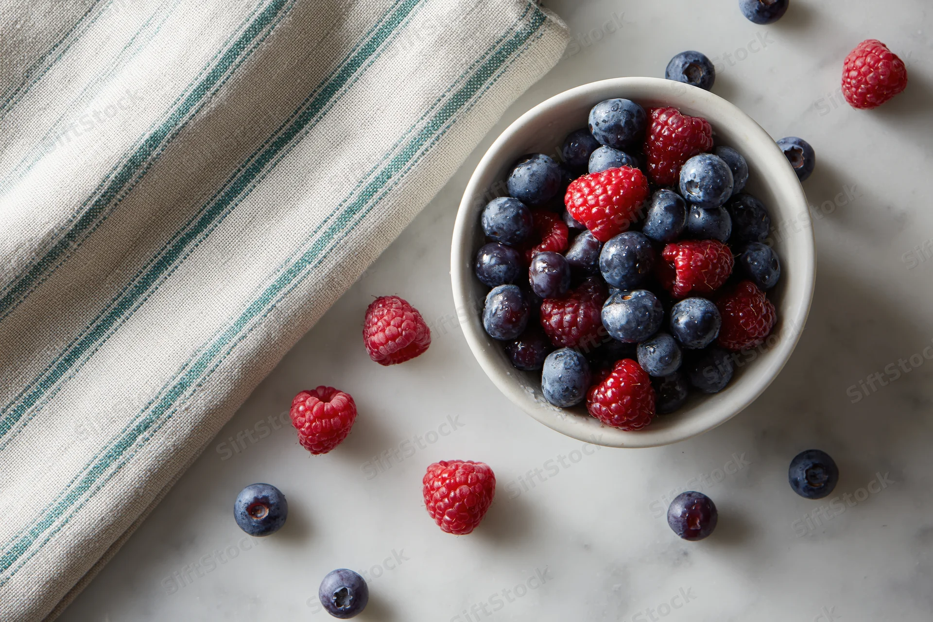 White ceramic bowl filled with fresh blueberries and raspberries on marble countertop beside striped cloth