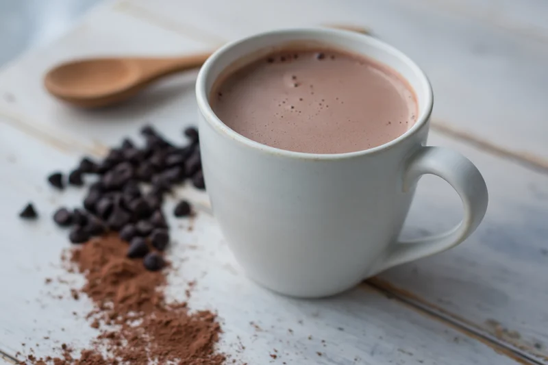 White ceramic mug filled with hot chocolate on a rustic wooden table beside cocoa powder and chocolate chips