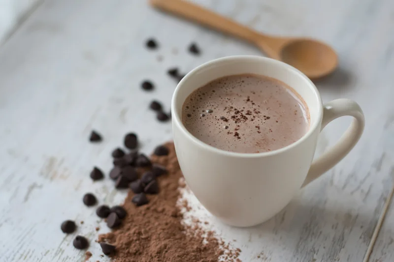 White ceramic mug filled with hot chocolate on a rustic wooden table with scattered chocolate chips and cocoa powder