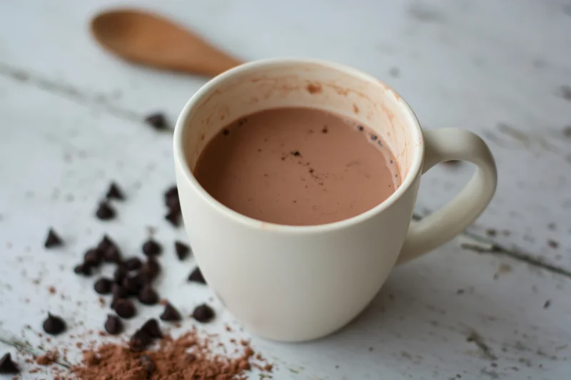 White ceramic mug filled with hot chocolate on a rustic wooden table with scattered chocolate chips and cocoa powder