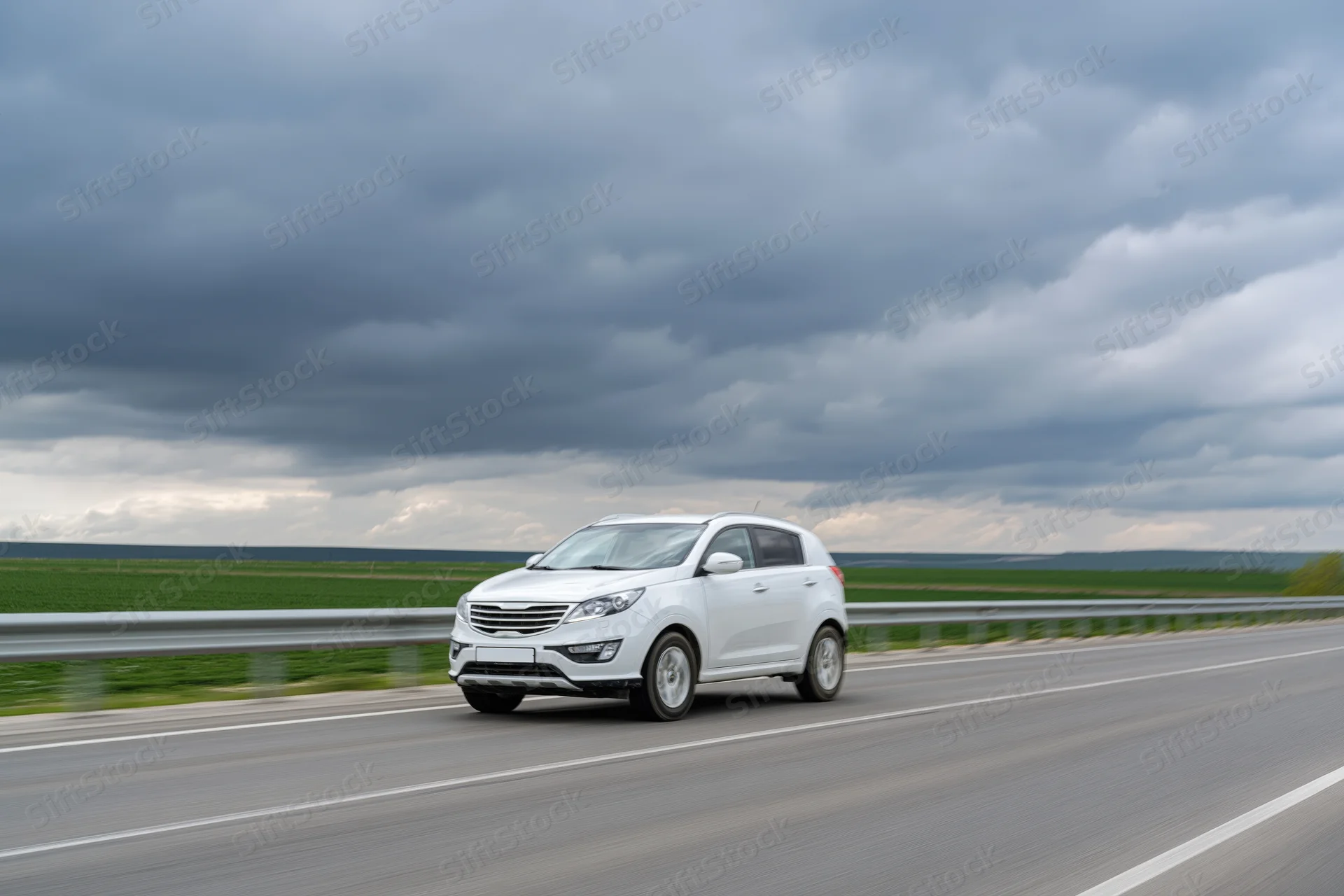 White compact suv driving on a two-lane highway with overcast sky and green fields in the background