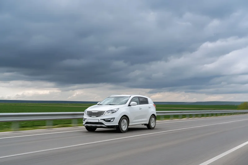 White compact suv driving on a two-lane highway with overcast sky and green fields in the background