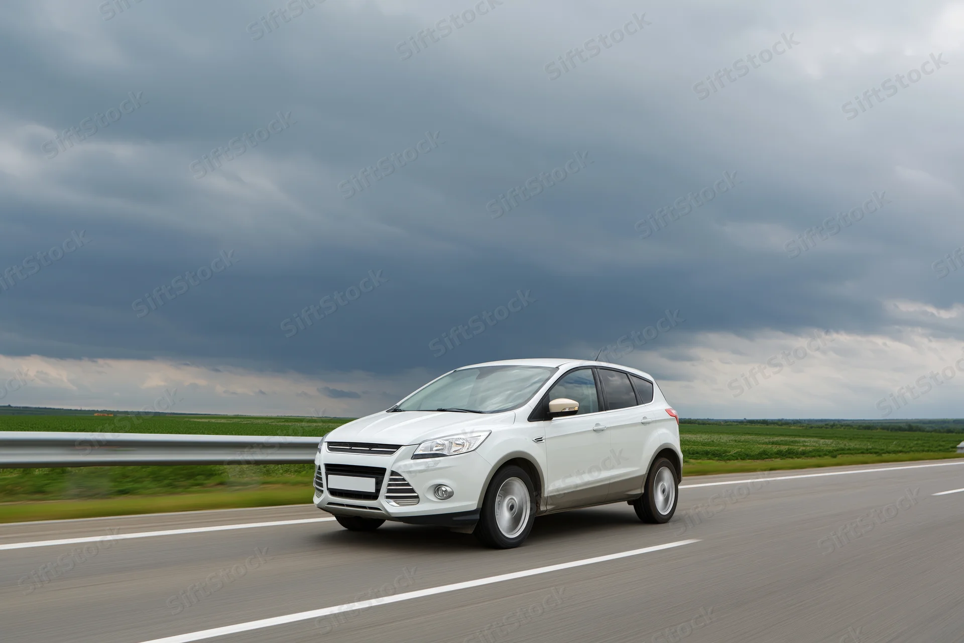 White compact suv driving on a two-lane highway with overcast sky and green fields alongside