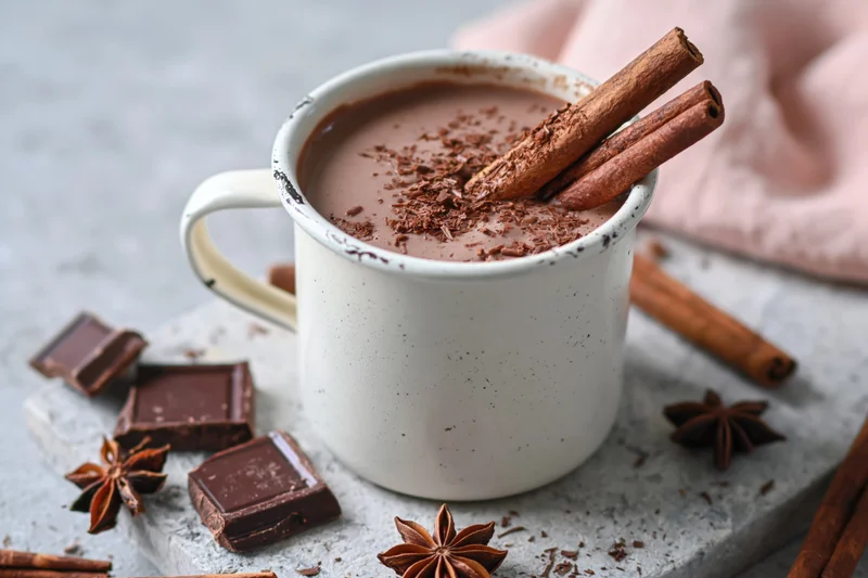 White enamel mug filled with hot chocolate topped with cinnamon sticks and chocolate shavings on a stone surface surrounded by chocolate pieces and star anise