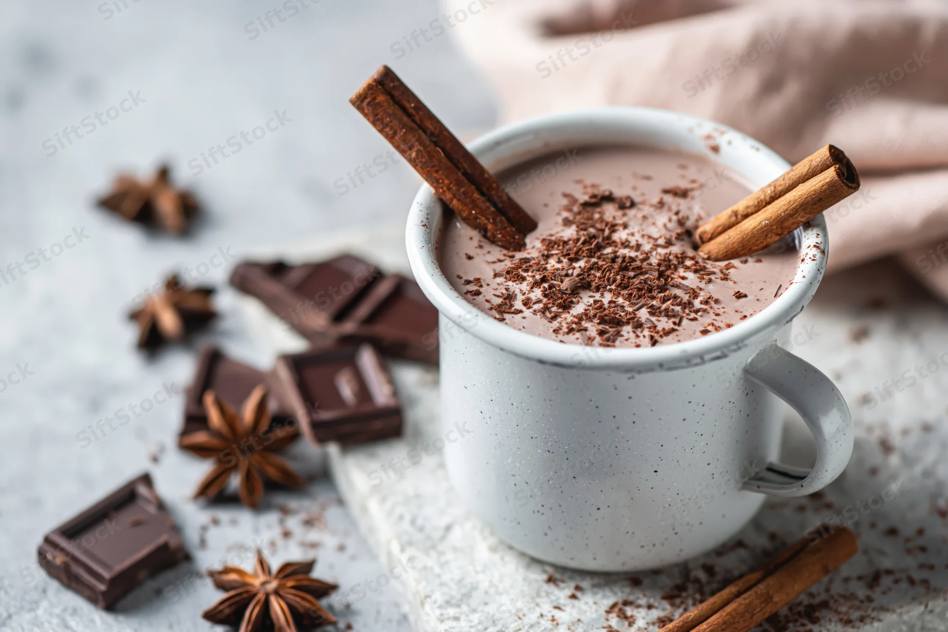 White speckled mug filled with hot chocolate topped with cinnamon sticks and chocolate shavings on a gray surface with scattered chocolate pieces and star anise