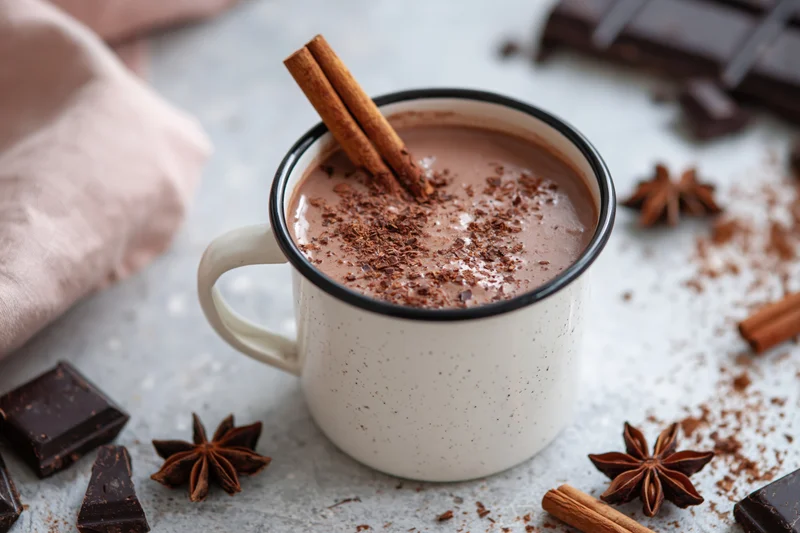 White enamel mug filled with hot chocolate topped with cinnamon sticks and cocoa powder on a textured surface surrounded by chocolate pieces and star anise