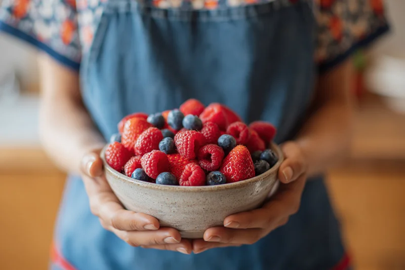 Woman holding a ceramic bowl filled with strawberries, raspberries and blueberries in a kitchen