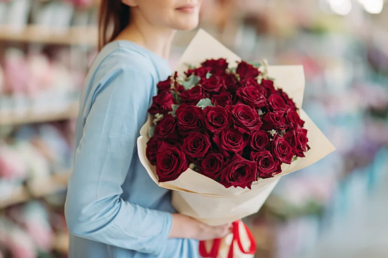 Woman holding large bouquet of deep red roses wrapped in paper in a flower shop