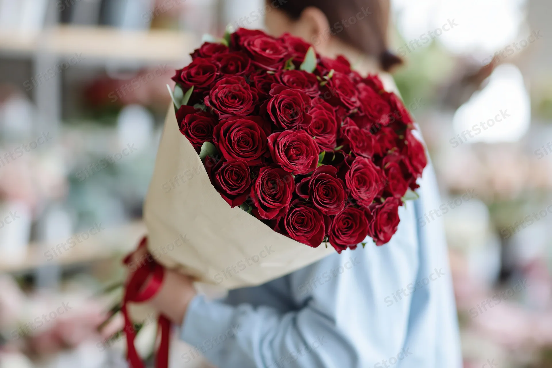 Woman holding large bouquet of deep red roses wrapped in paper with ribbon, in a flower shop setting