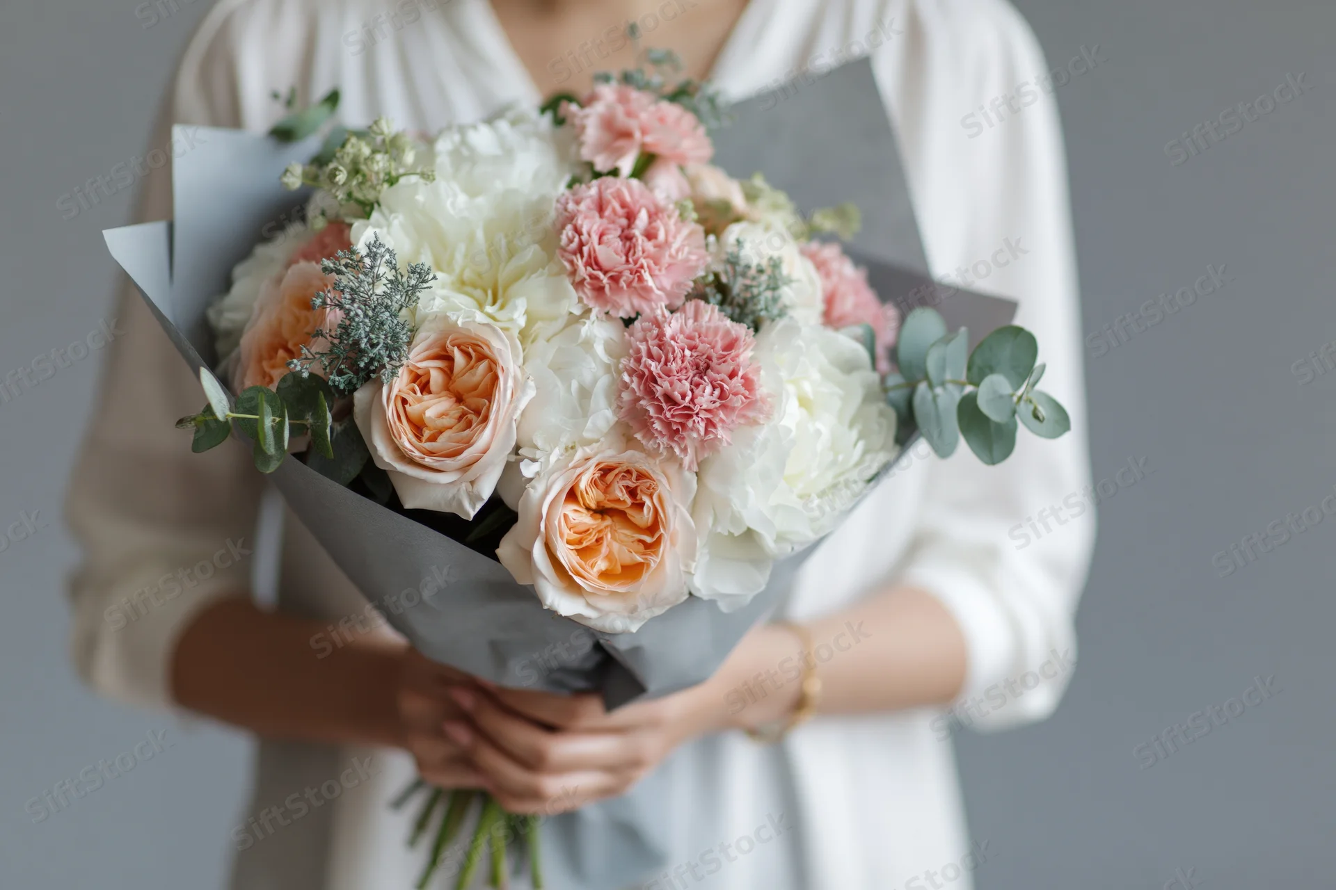 Woman holding a large wrapped bouquet of peach and white roses with greenery against a gray background