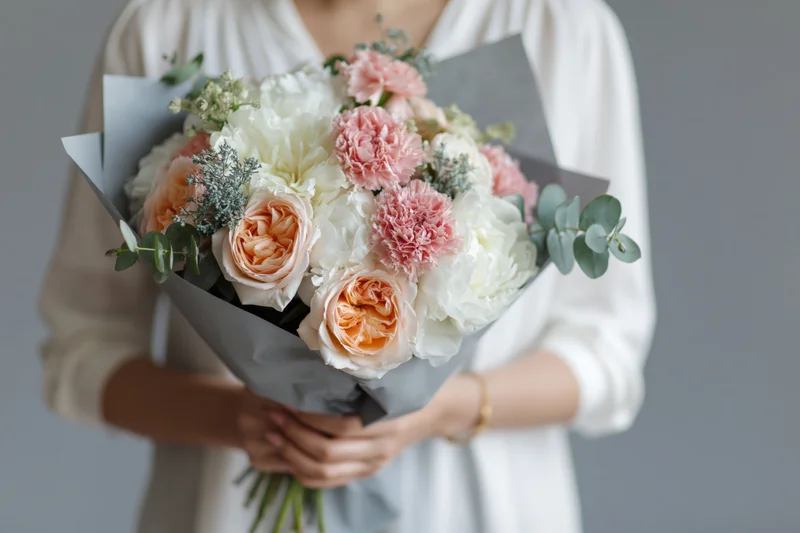 Woman holding a large wrapped bouquet of peach and white roses with greenery against a gray background