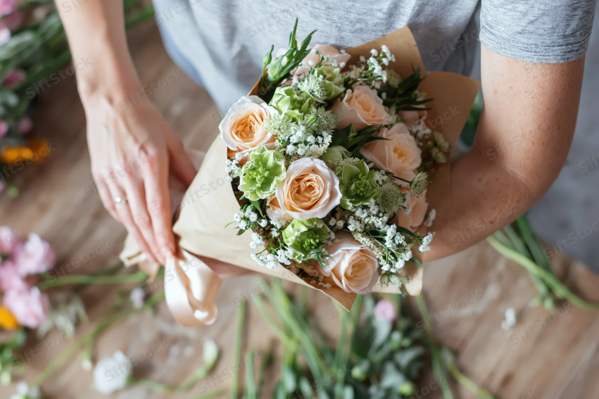 Woman wrapping a bouquet of roses and greenery with paper and ribbon on a wooden table