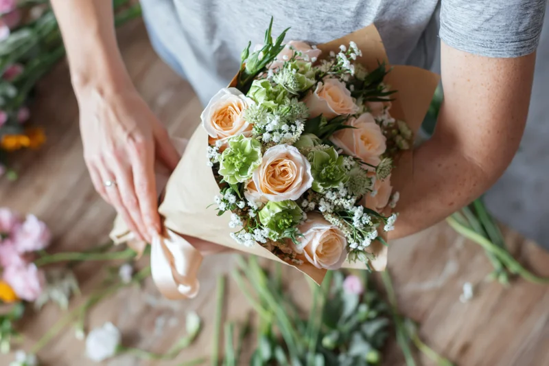 Woman wrapping a bouquet of roses and greenery with paper and ribbon on a wooden table