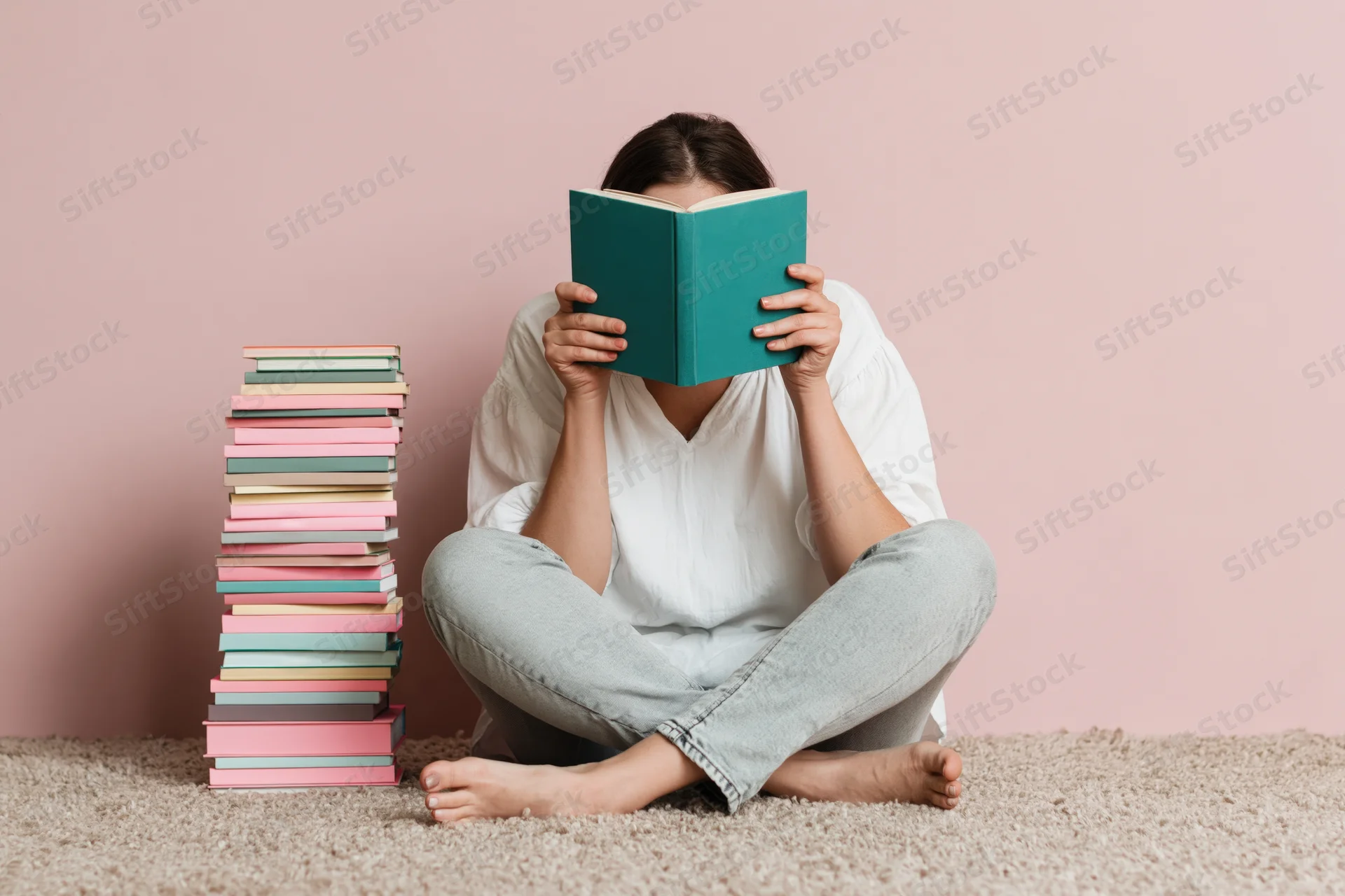 Woman sitting cross-legged on carpet reading book with stack of books beside her