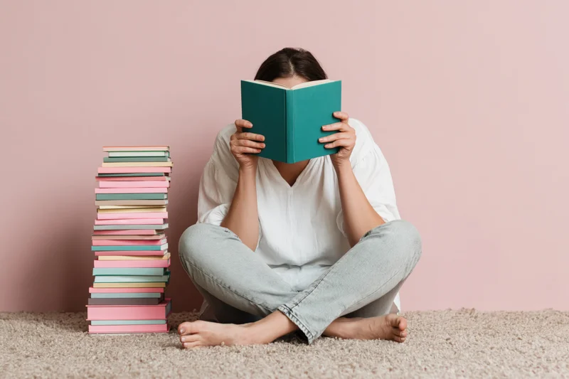 Woman sitting cross-legged on carpet reading book with stack of books beside her