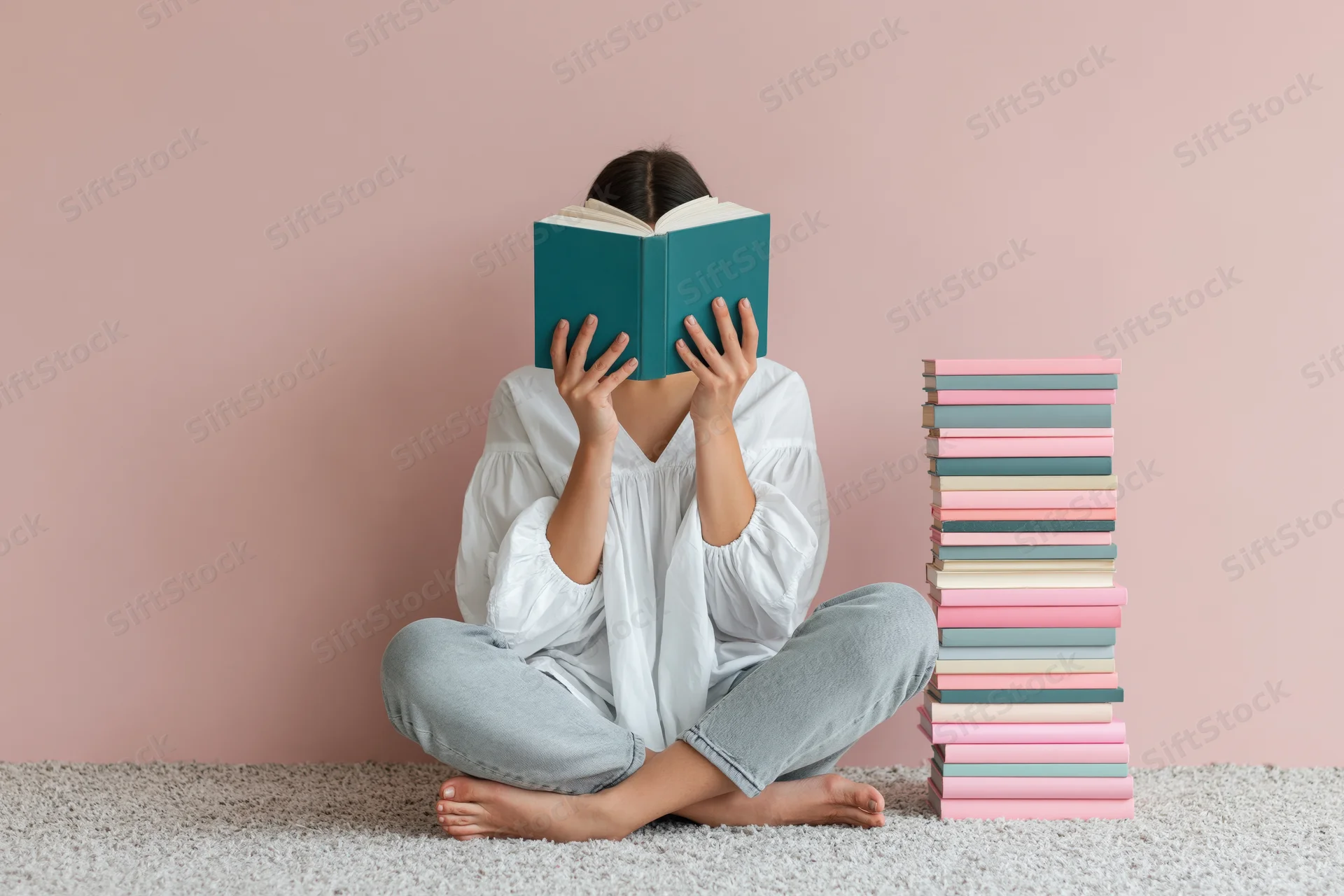 Woman sitting cross-legged on carpet reading book with stack of books beside her