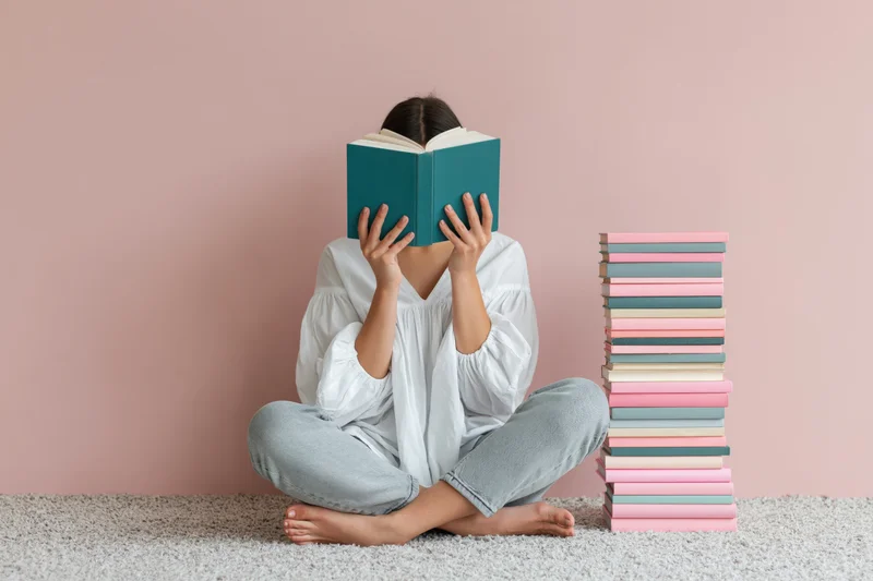 Woman sitting cross-legged on carpet reading book with stack of books beside her