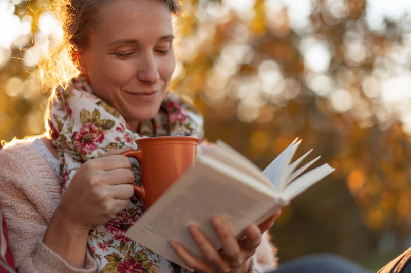 Woman reading a book while holding a mug outdoors in autumn sunlight