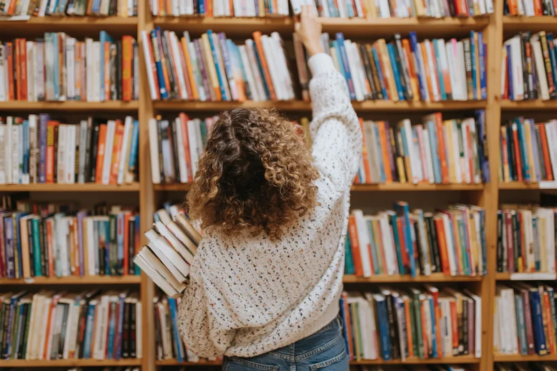 Woman reaching for a book on a high shelf in a library while holding several books in her other arm