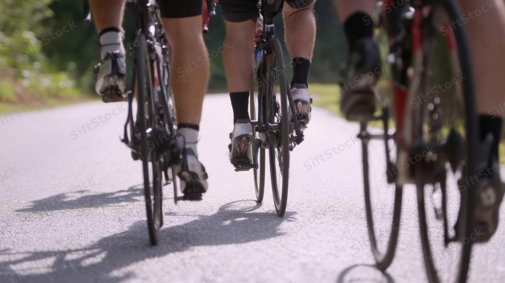 Cyclists pedaling in a group on a paved road, viewed from behind focusing on legs and shoes clipped into pedals
