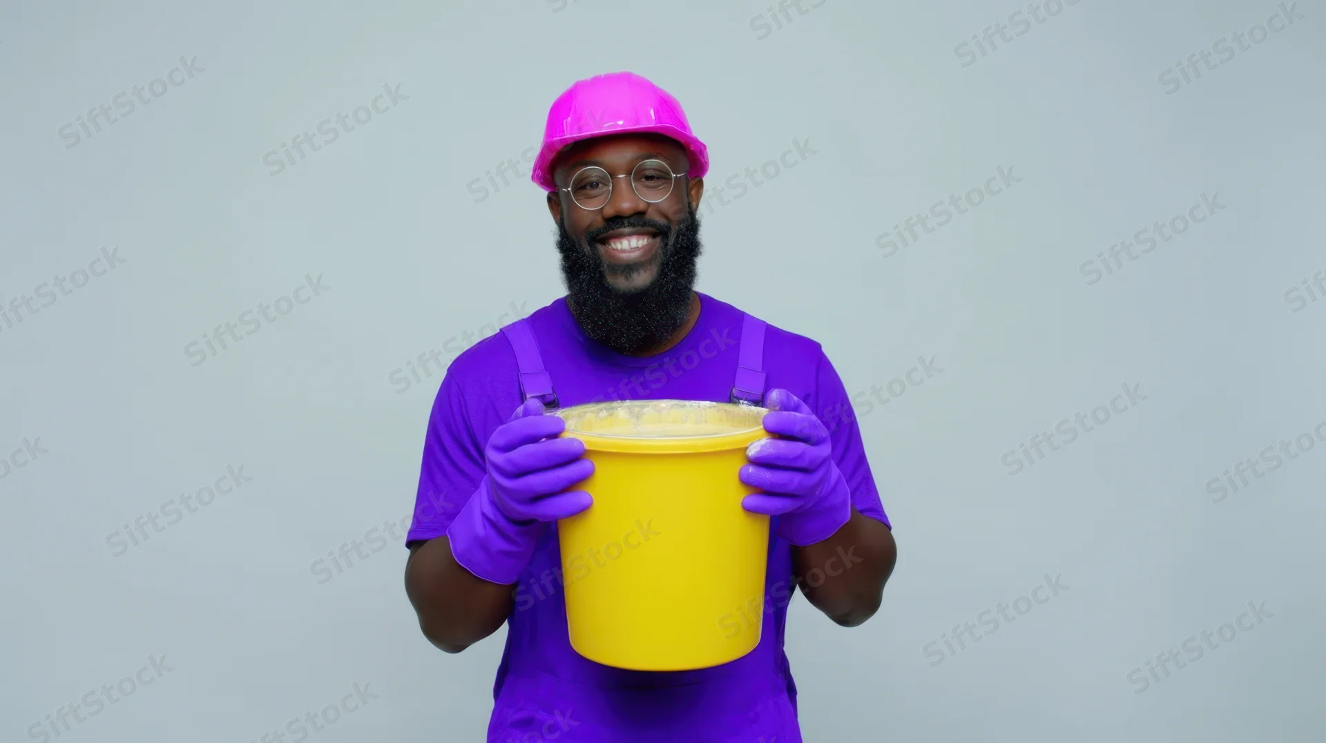 Smiling man in purple overalls and pink hard hat holding yellow bucket against plain background