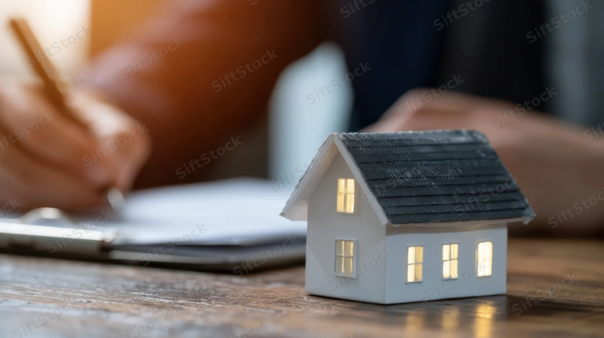 Miniature house model with glowing windows on wooden desk beside hands writing on document