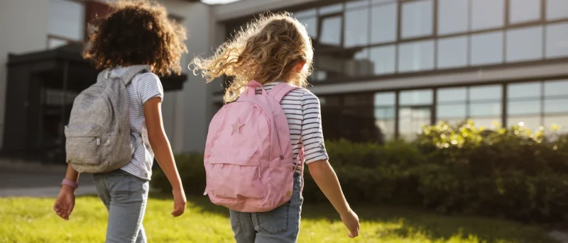 Two young girls with backpacks walking toward a school building on a sunny day