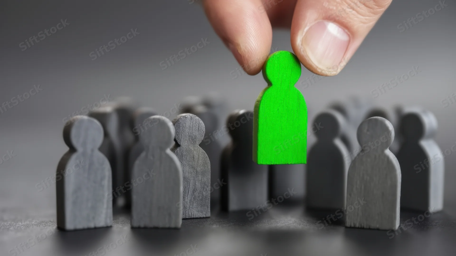 Hand lifting a green wooden figure from a group of gray figures on a dark surface