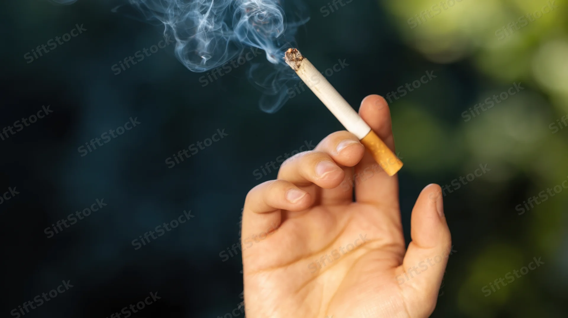 Hand holding a lit cigarette with smoke rising against a blurred green outdoor background