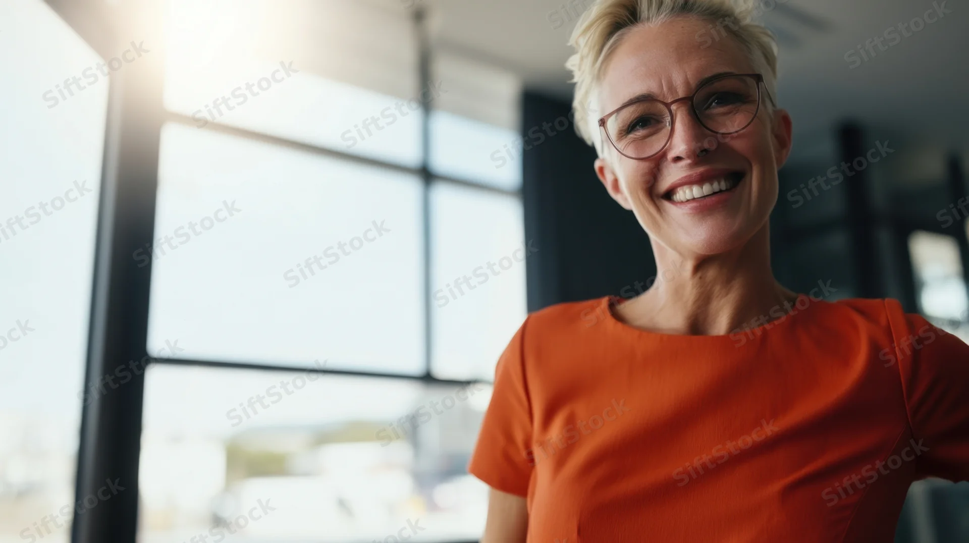 Smiling woman in orange blouse wearing glasses standing in modern office near window
