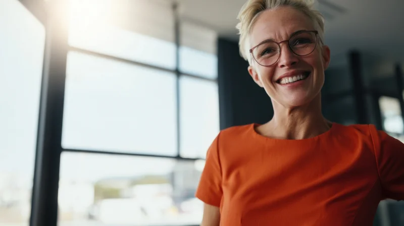 Smiling woman in orange blouse wearing glasses standing in modern office near window