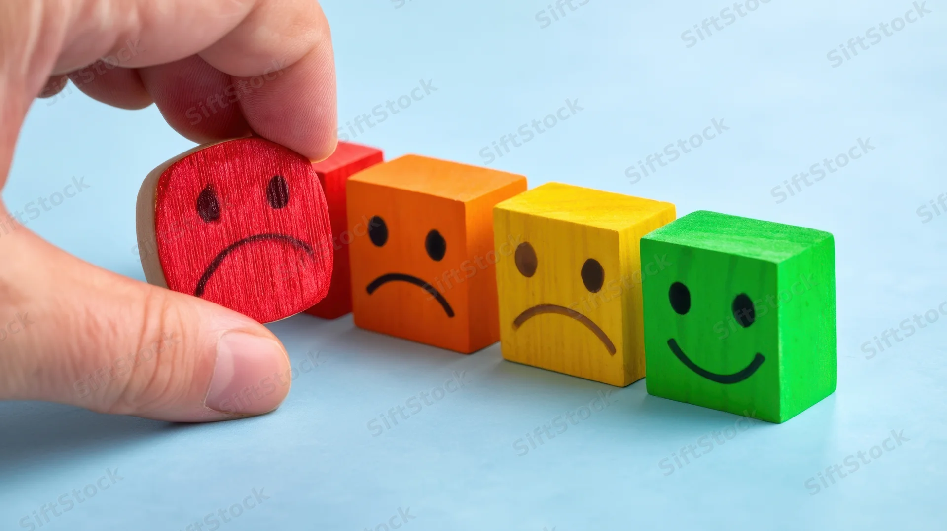 Hand selecting a red sad face block from a row of colored emotion cubes on a blue surface