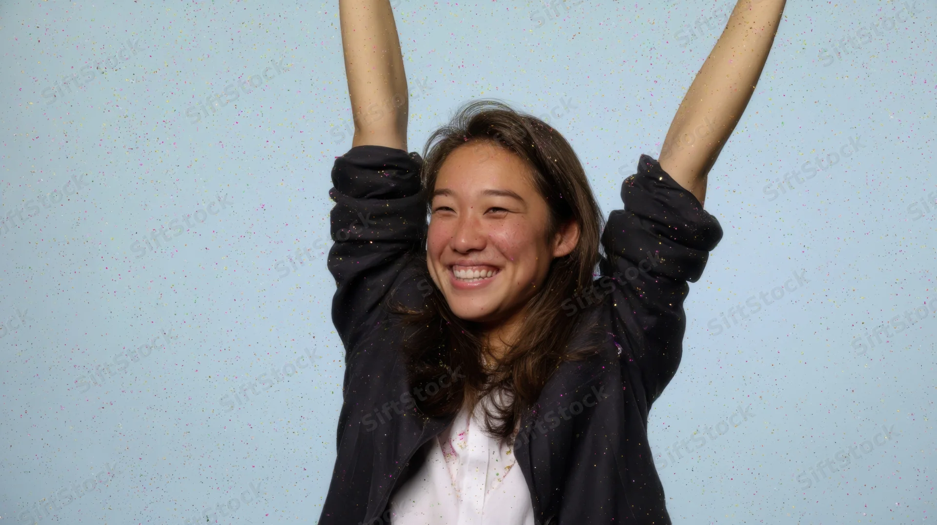 Young woman smiling with arms raised amidst falling confetti against blue background
