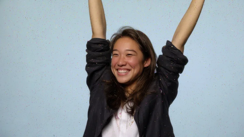 Young woman smiling with arms raised amidst falling confetti against blue background