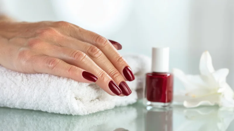 Woman's hand with red manicure resting on white towel beside nail polish bottle and flower