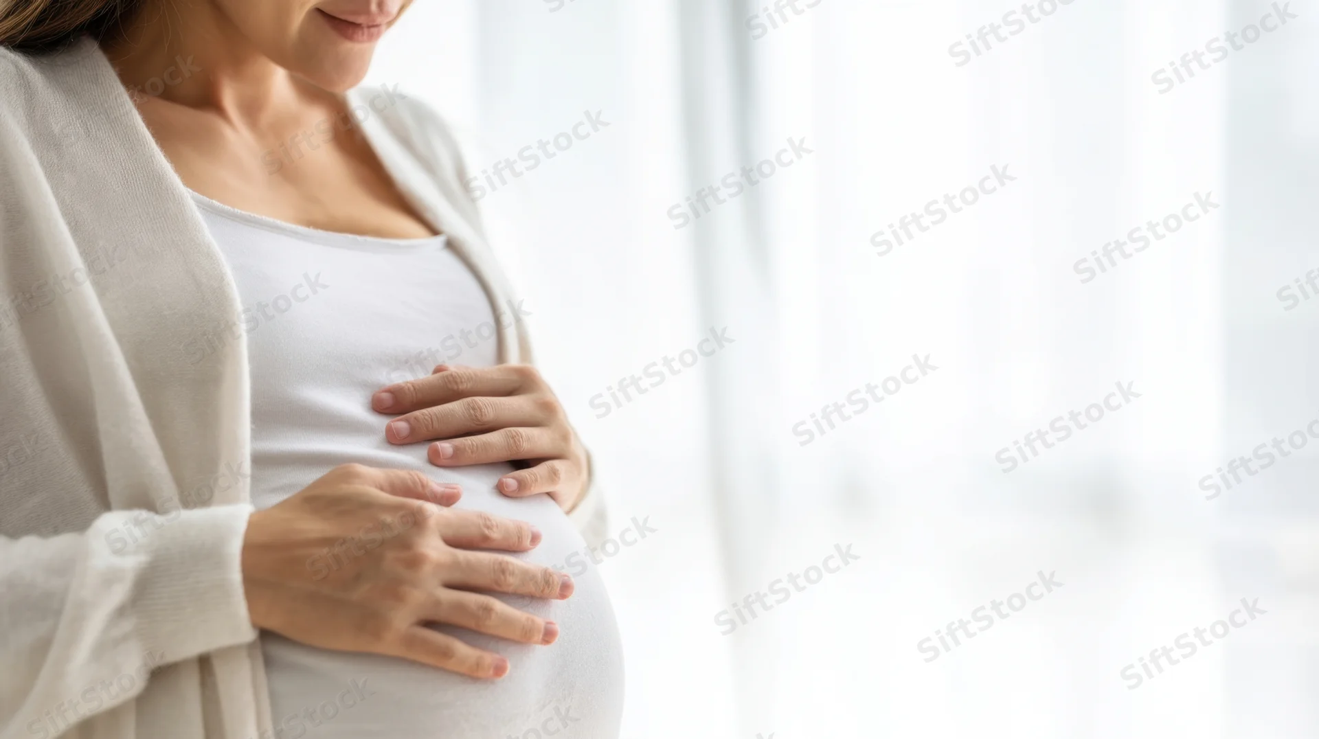 Pregnant woman cradling her belly indoors by a window