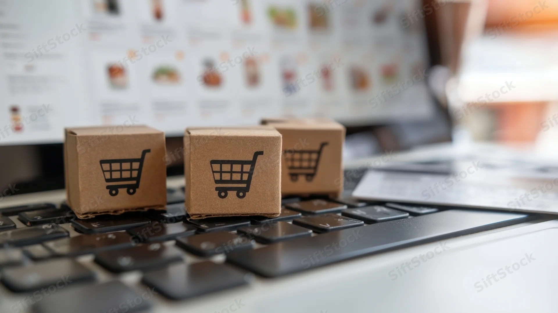 Small cardboard boxes with shopping cart symbols placed on laptop keyboard in front of online store screen