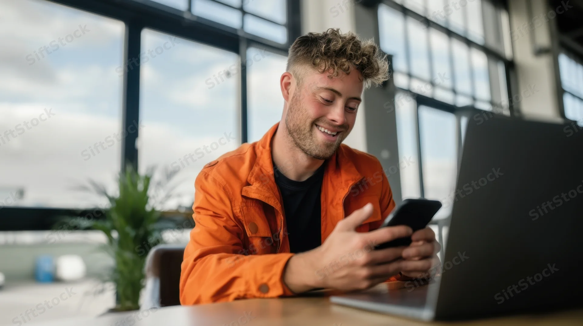 Young man smiling while using phone at desk with laptop in modern office
