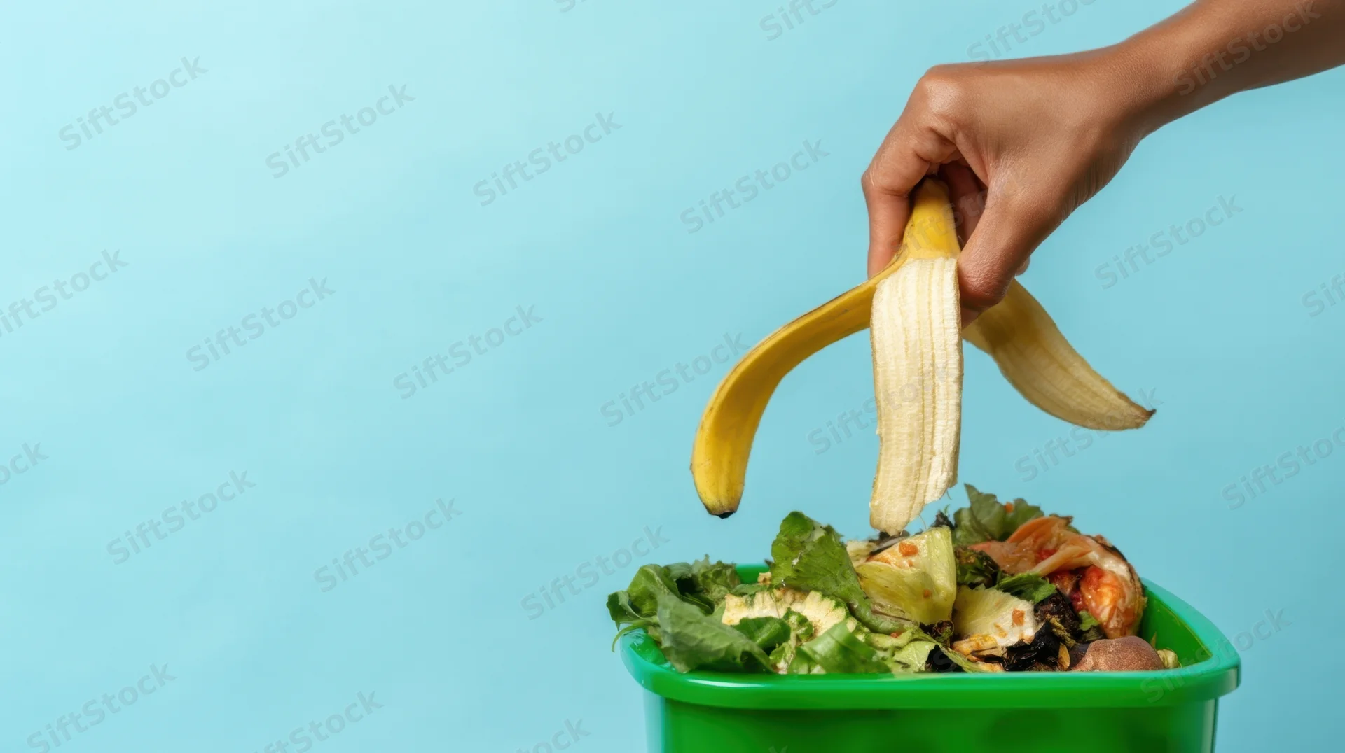 Hand dropping banana peel into green compost bin against blue background