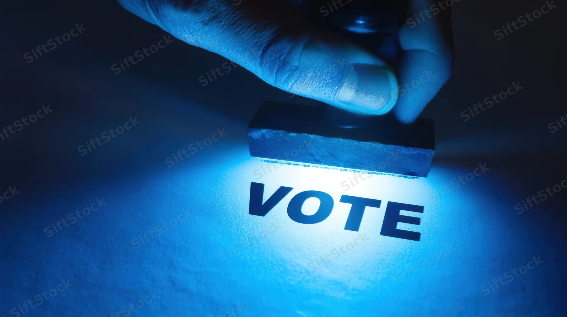 Hand pressing rubber stamp onto paper to mark vote in blue light